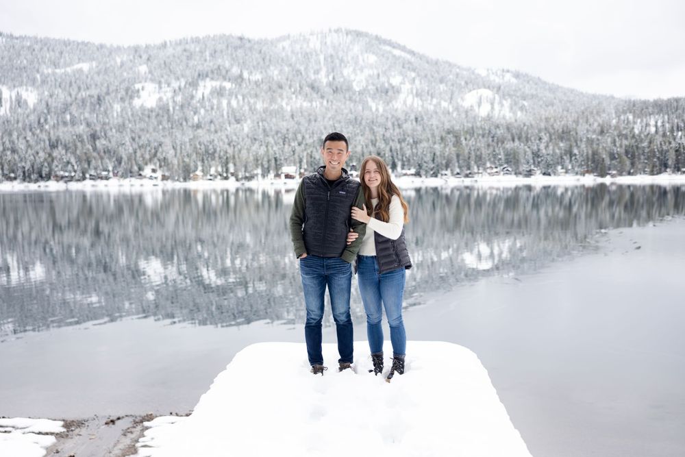 A couple stands on a snowy ledge overlooking a serene winter lake surrounded by snow-capped mountains.