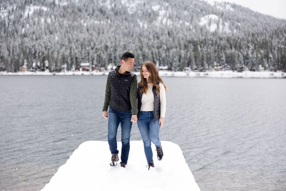 A couple stands close together on a snow-covered dock overlooking a serene mountain lake in winter.