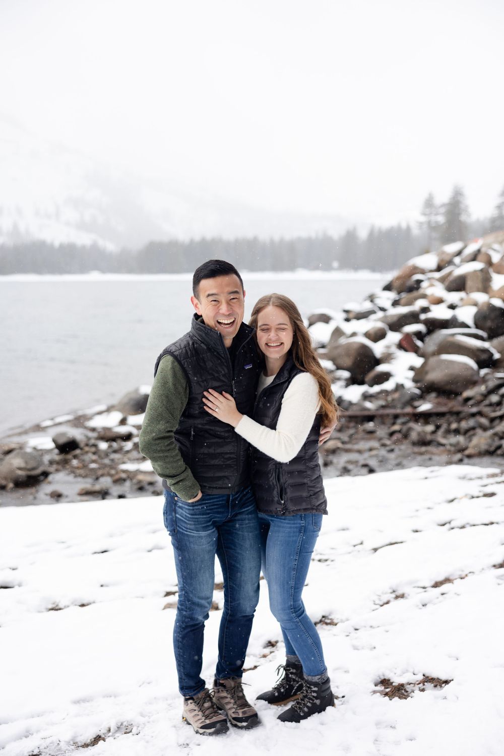 A couple embraces while standing on a snowy shoreline with rocky water's edge and misty mountains in the background.