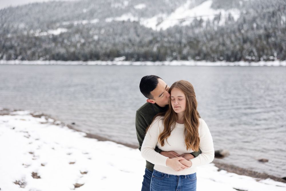 A couple embraces by a snowy mountain lake with evergreen trees in winter.