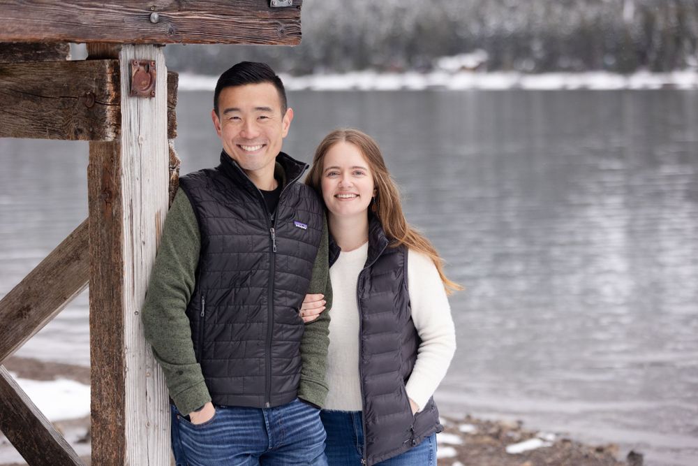 A couple in matching black puffer vests poses together by a lake during winter.