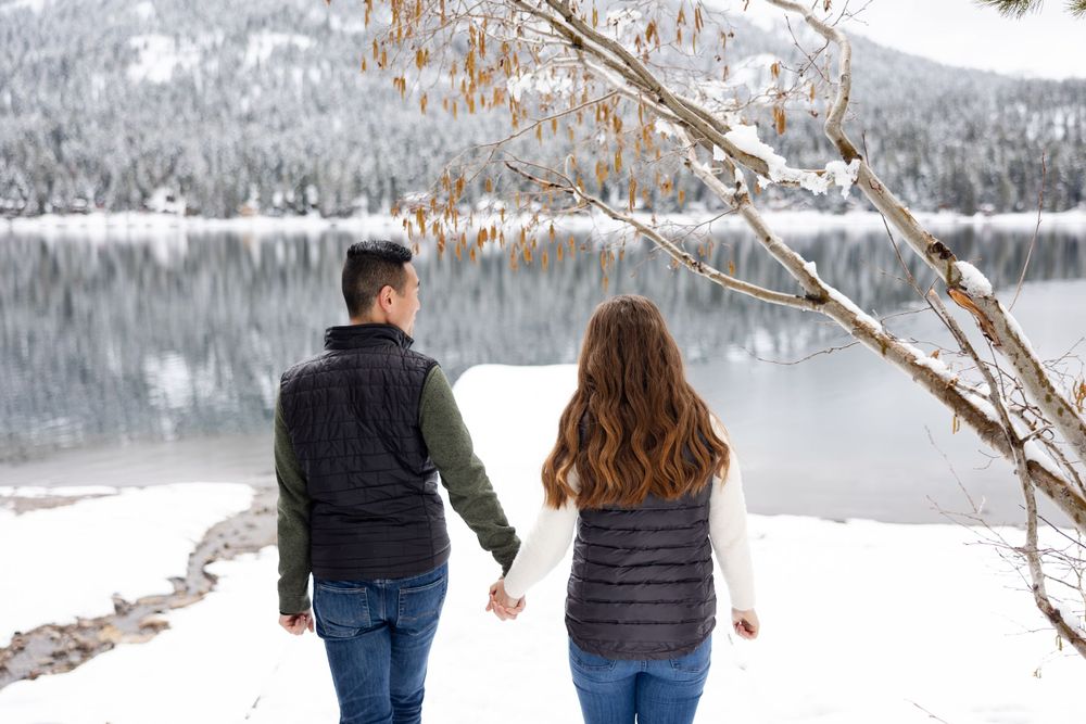 Two people in dark winter jackets holding hands while walking along a snowy lakeshore beneath bare branches.