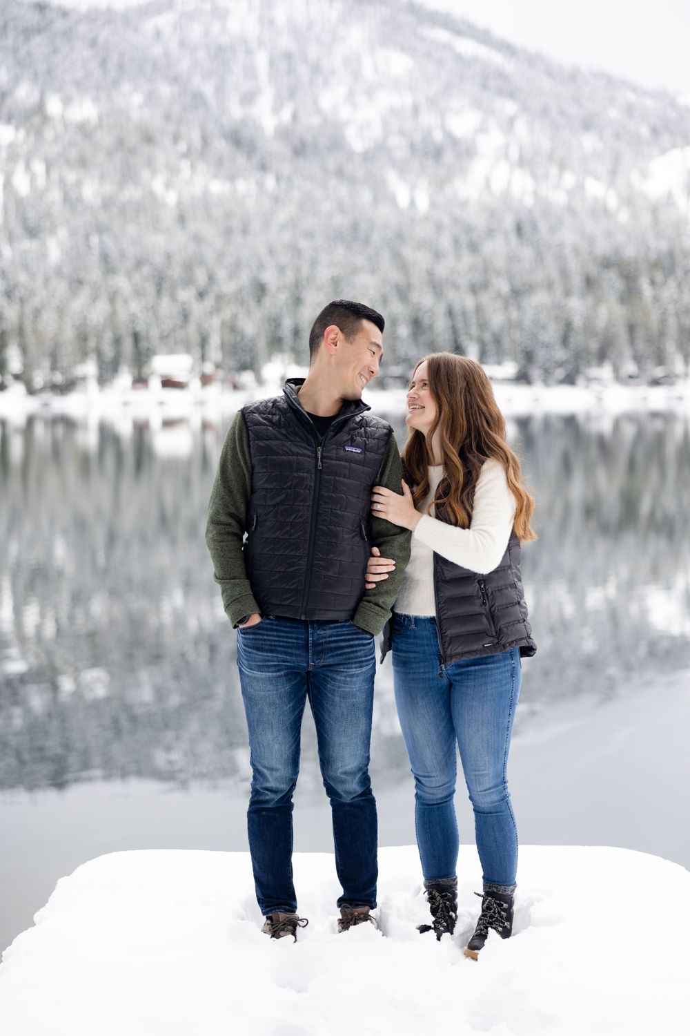 A couple wearing matching winter vests and jeans stands together in the snow near the serene Donner Lake.