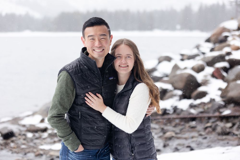 A happy couple embraces while posing for a winter portrait by a snowy rocky shoreline.