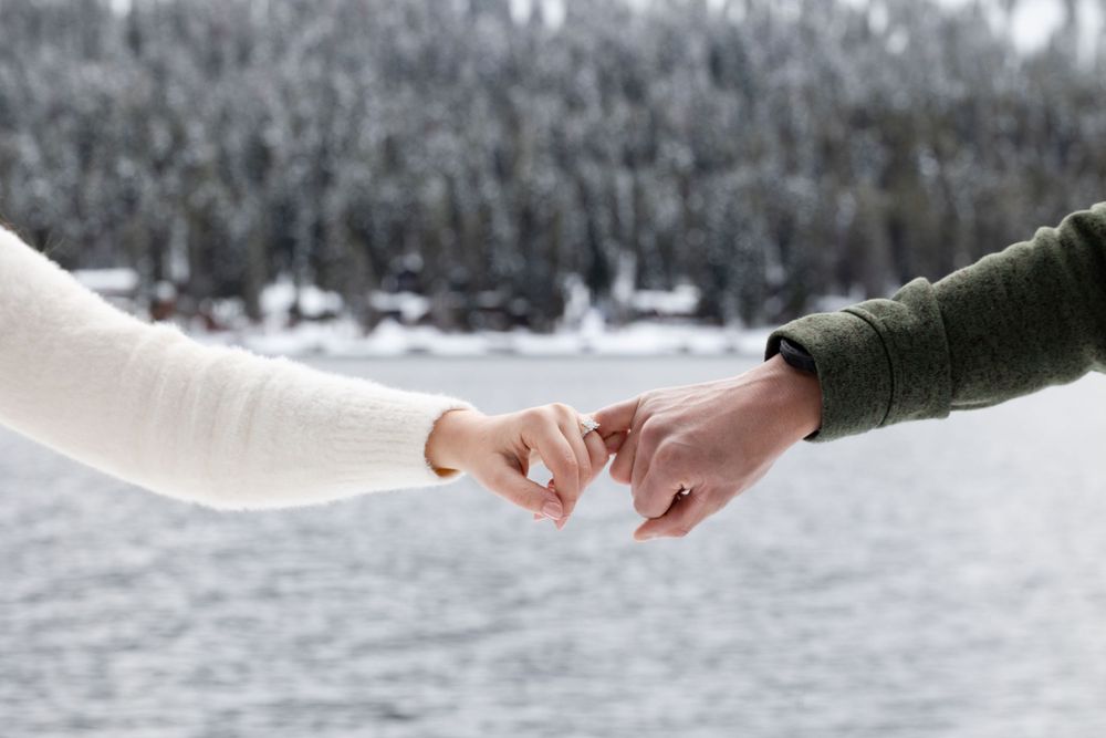 Two hands reaching out to connect with pinky fingers against a snowy winter landscape with forest in the background.
