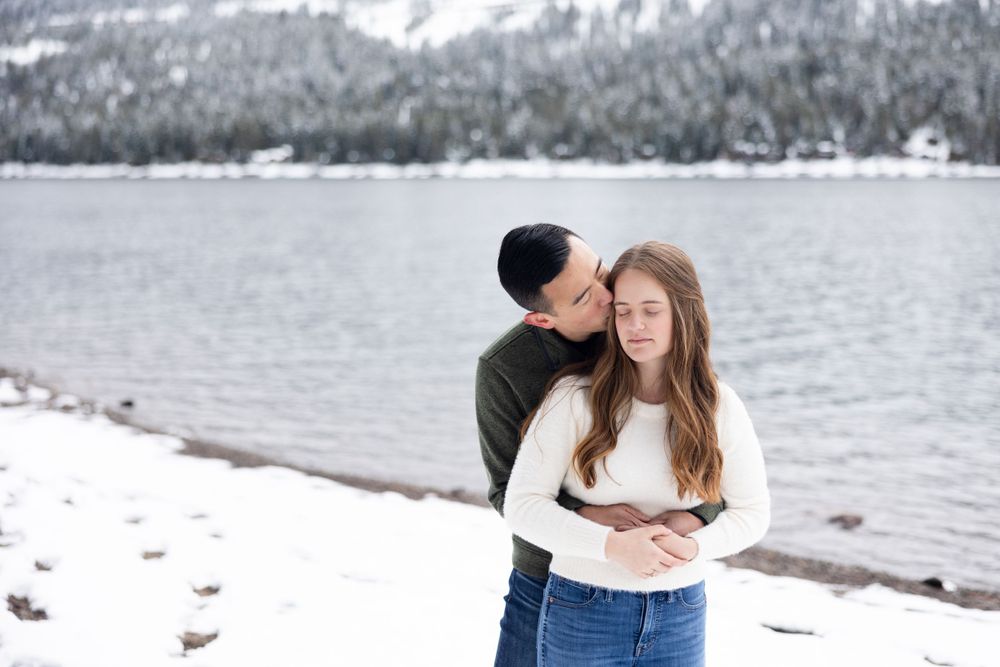 A romantic winter scene with a couple embracing by a snowy lake with mountain views in the background.