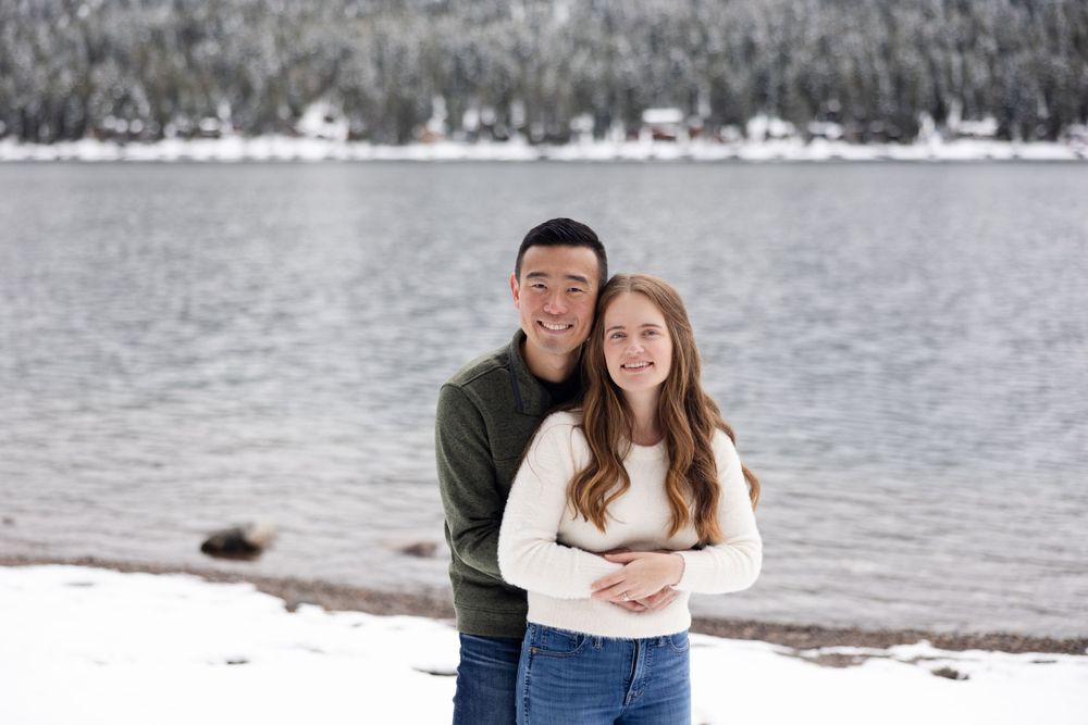 A couple embraces while standing on a snowy lakeshore with mountains and trees visible in the background.