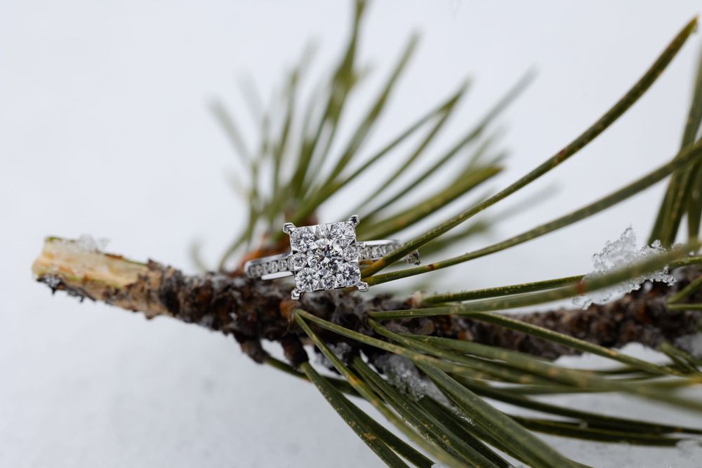 Diamond engagement ring with flower-shaped setting perched on snowy pine branch against white background.