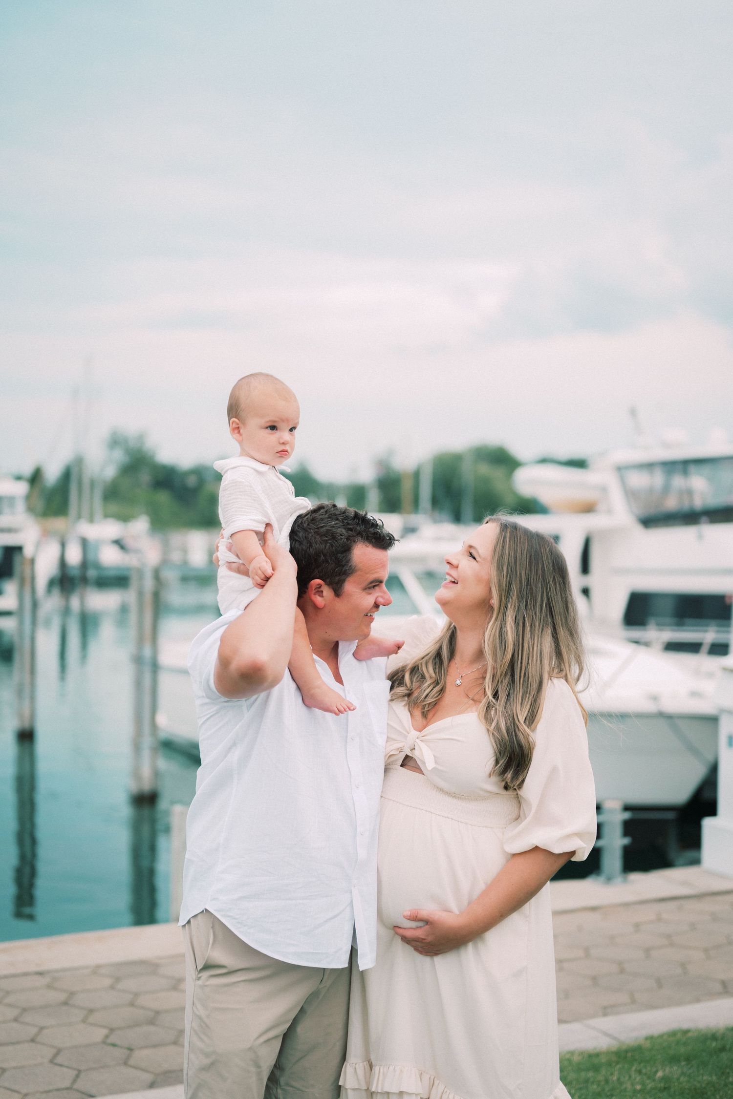 A happy couple at a marina expecting a baby share a tender moment with their toddler on dad's shoulders.