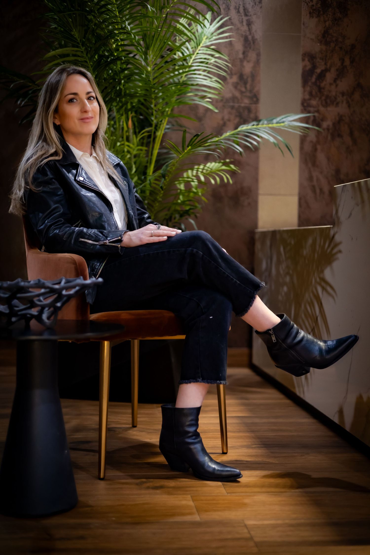A person in a leather jacket and boots sits casually on a chair next to a palm plant in a modern interior setting.