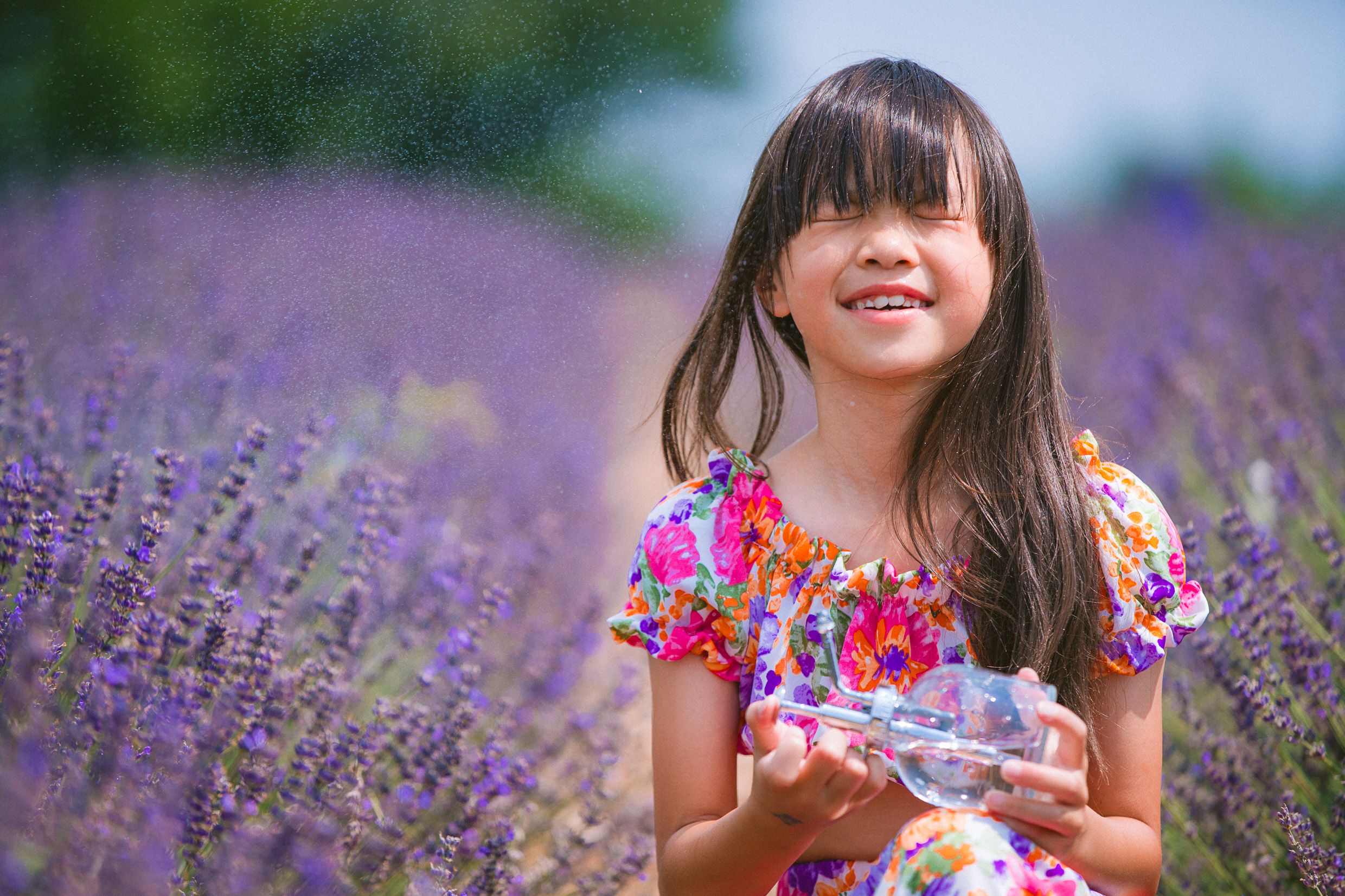 Child in a floral dress holds a glass jar while sitting among blooming purple lavender flowers in a summer field.