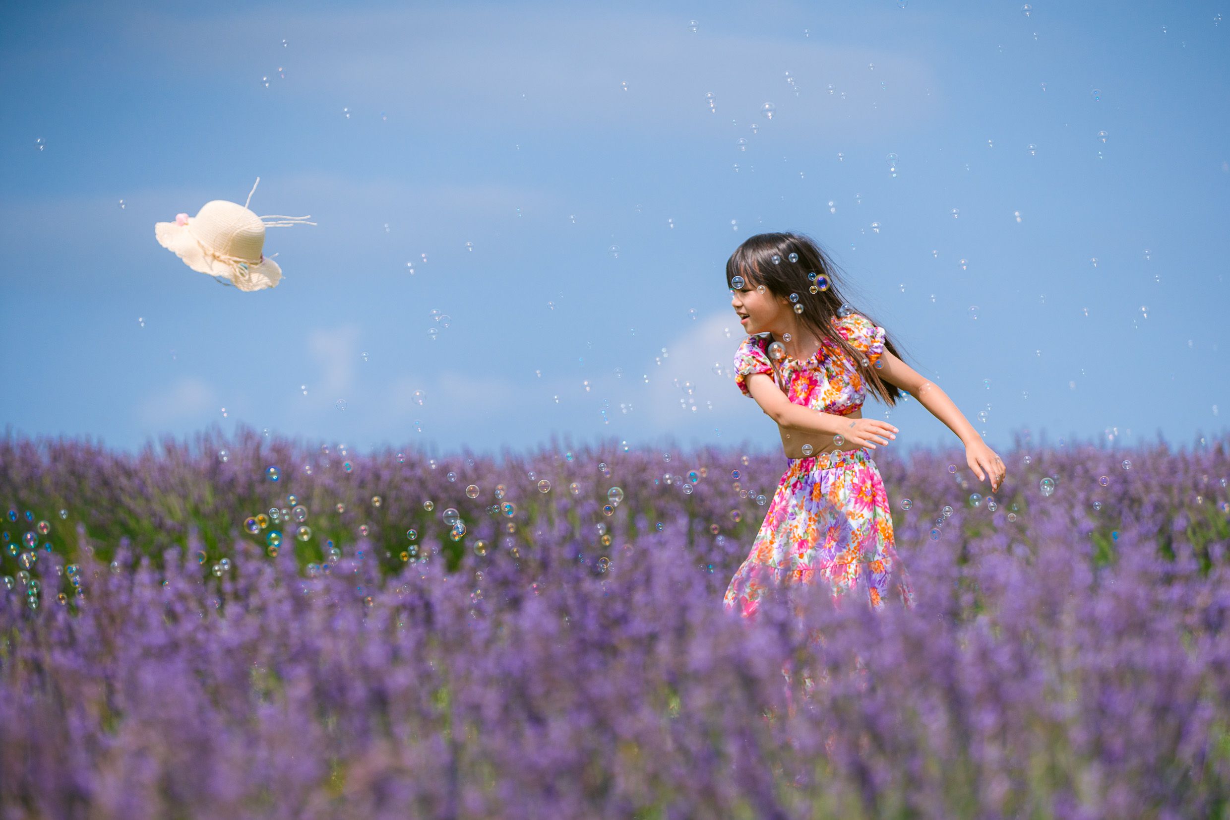 A figure in a floral dress tosses a hat while standing in a vibrant purple lavender field under blue summer skies.