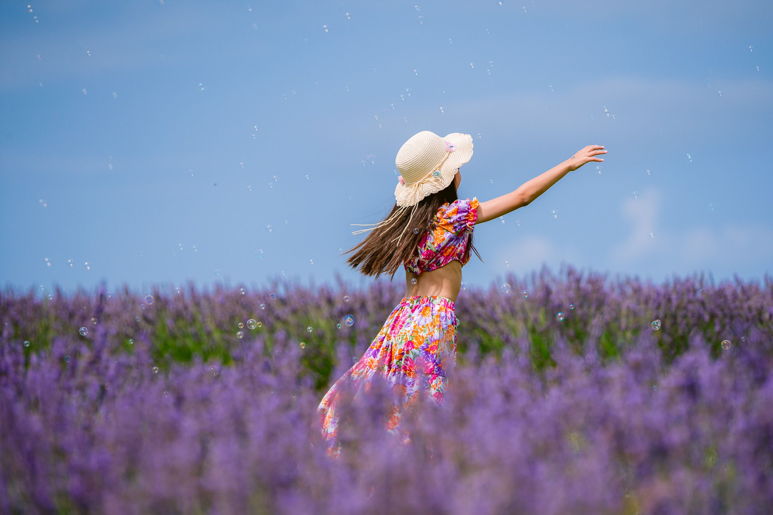 A person in a floral dress and sun hat dances joyfully in a blooming purple lavender field against a bright blue sky.