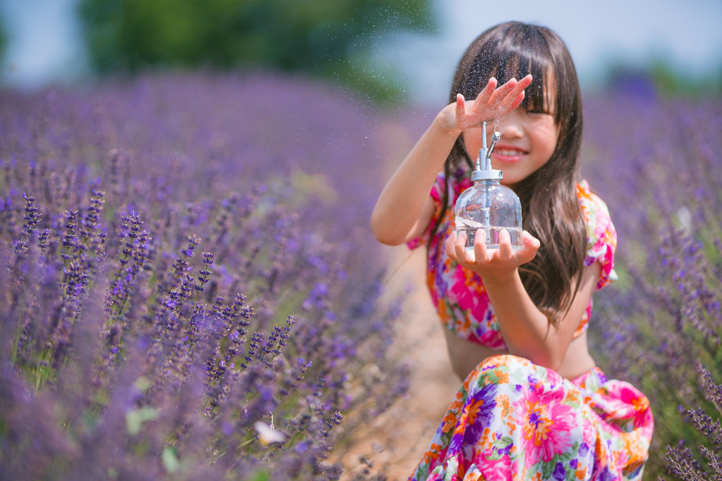 A child in a colorful floral dress holds a small bottle while standing in a blooming lavender field on a summer day.