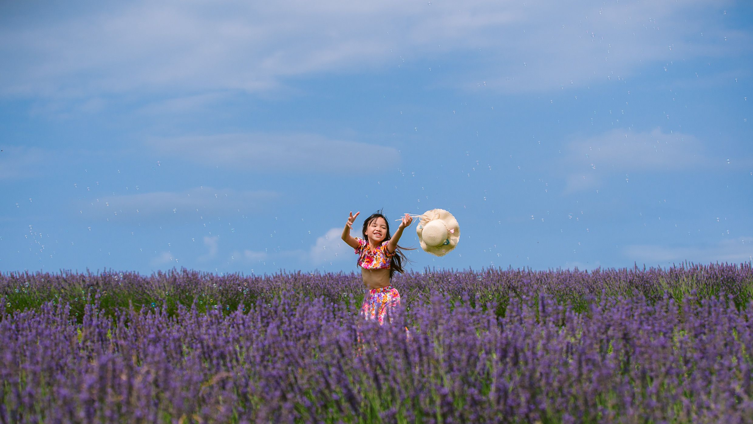 Someone jumps joyfully with a straw hat in a vibrant purple lavender field against a blue summer sky.