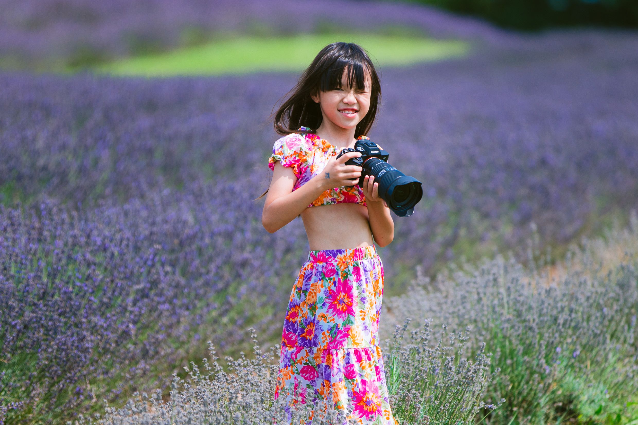 A colorfully dressed figure holds a camera while standing in a blooming purple lavender field on a summer day.