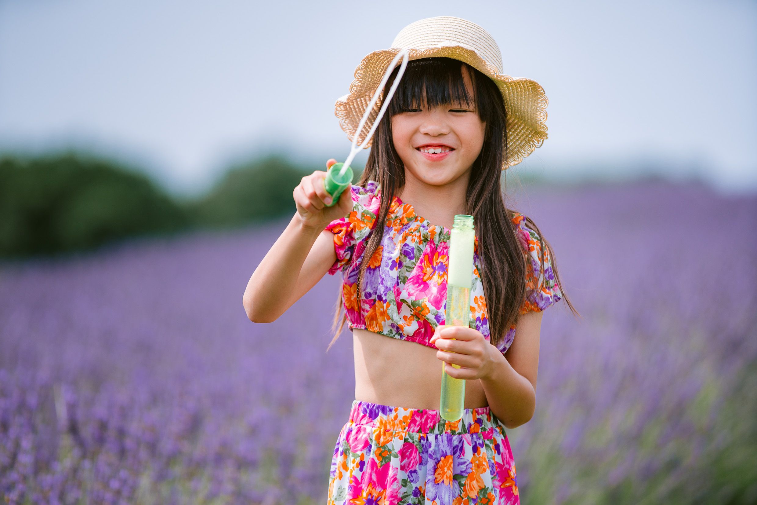 A figure wearing a floral dress and straw hat stands in a blooming lavender field against a blue summer sky.