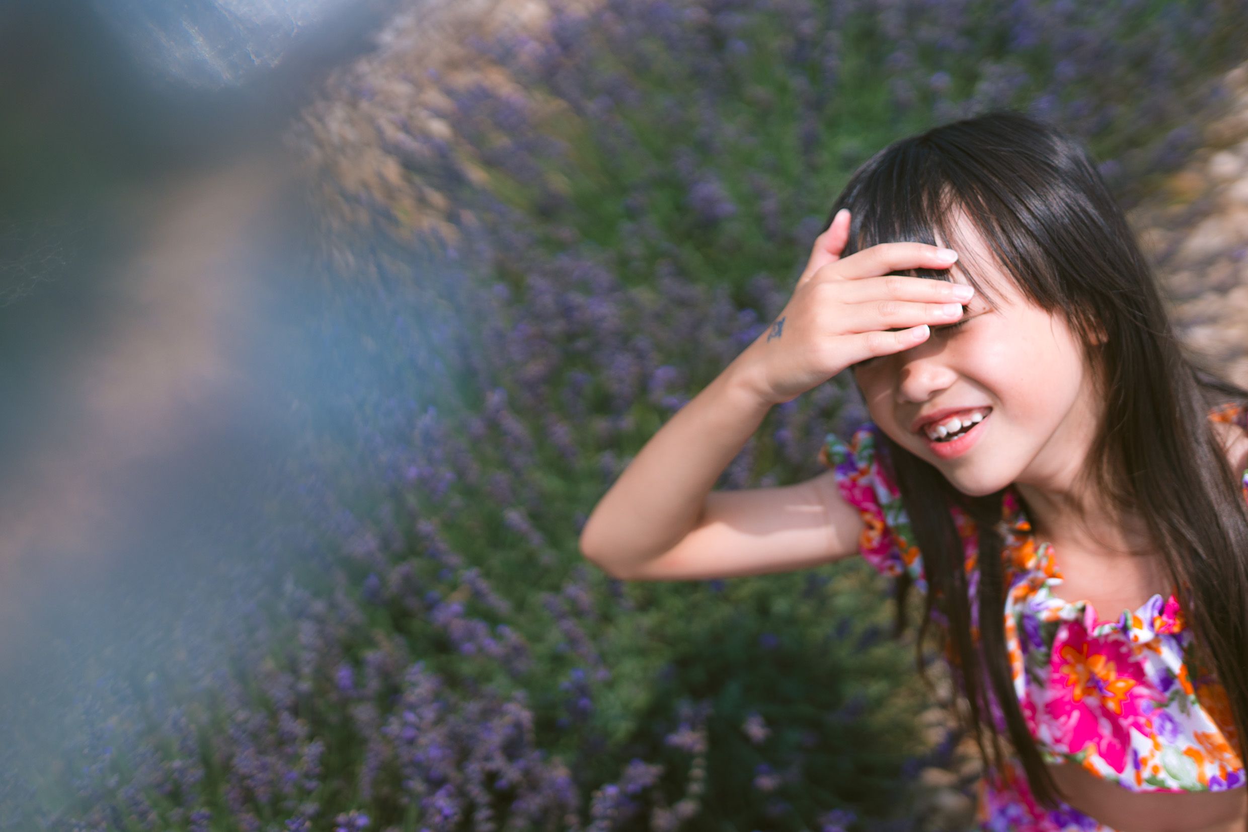 A person in a floral dress laughs while covering their face, standing among purple blooming flowers on a sunny day.