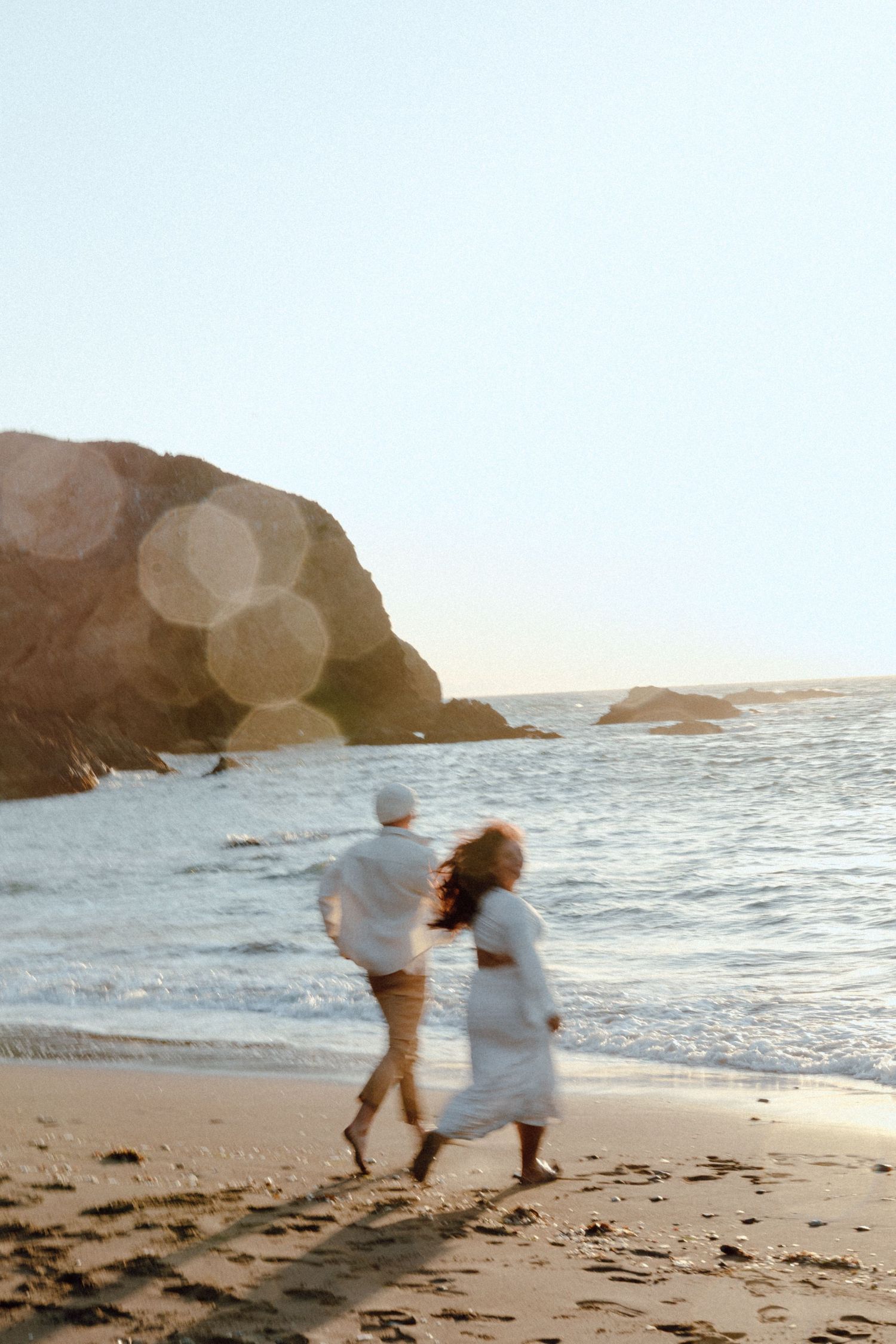 A couple in white clothing runs joyfully along a scenic beach at sunset with cliffs and ocean waves in the background.