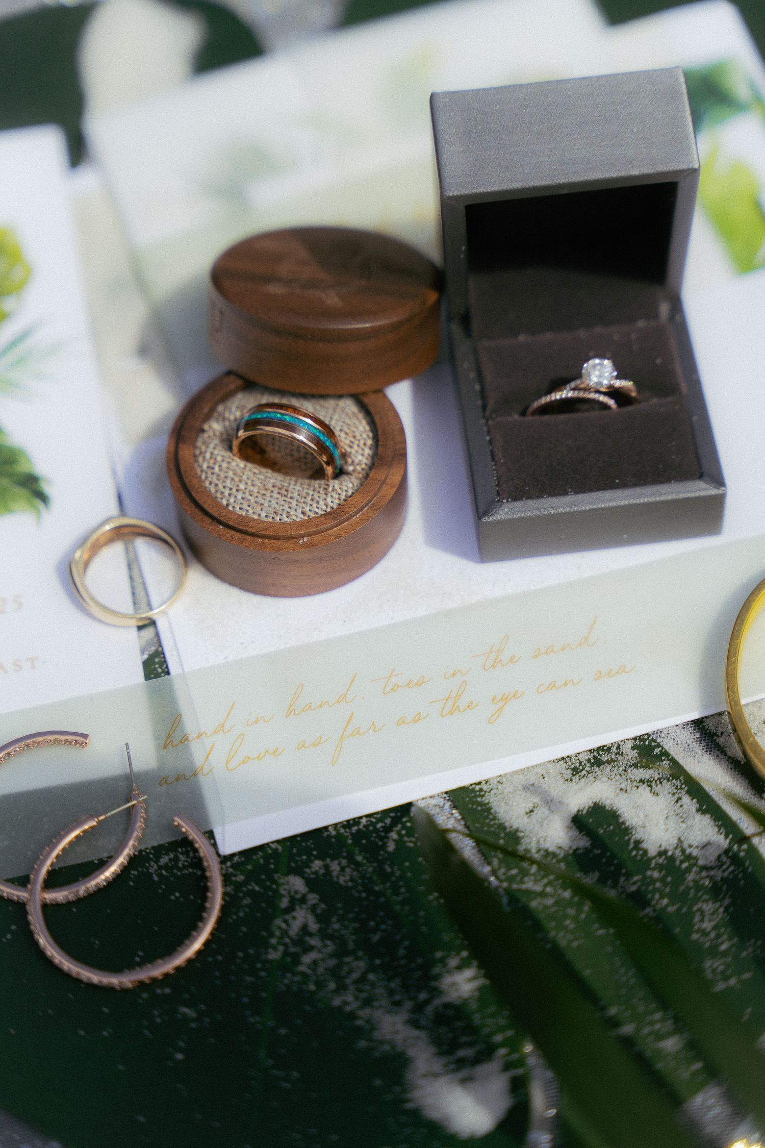 Wedding rings and jewelry displayed in wooden and black boxes on a tropical leaf background.