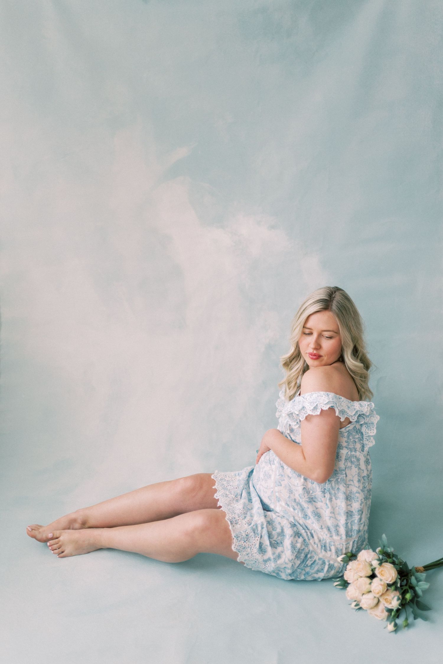 Soft ethereal maternity photoshoot with light floral dress and pink roses against powder blue backdrop.