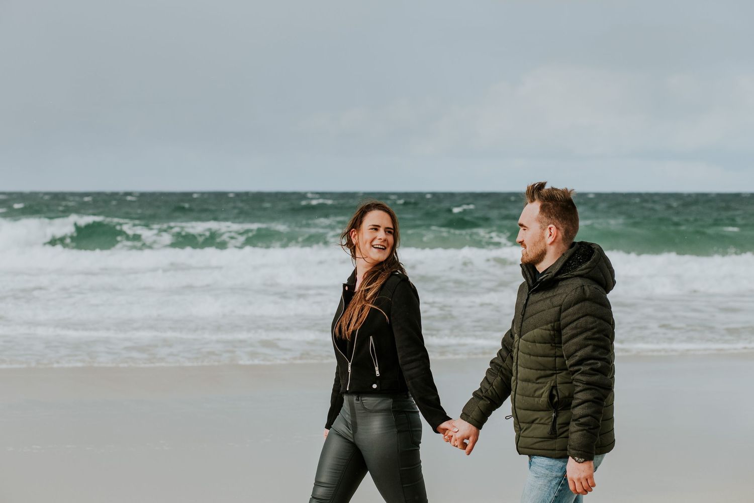 A couple holds hands while enjoying waves crashing on a windy beach.
