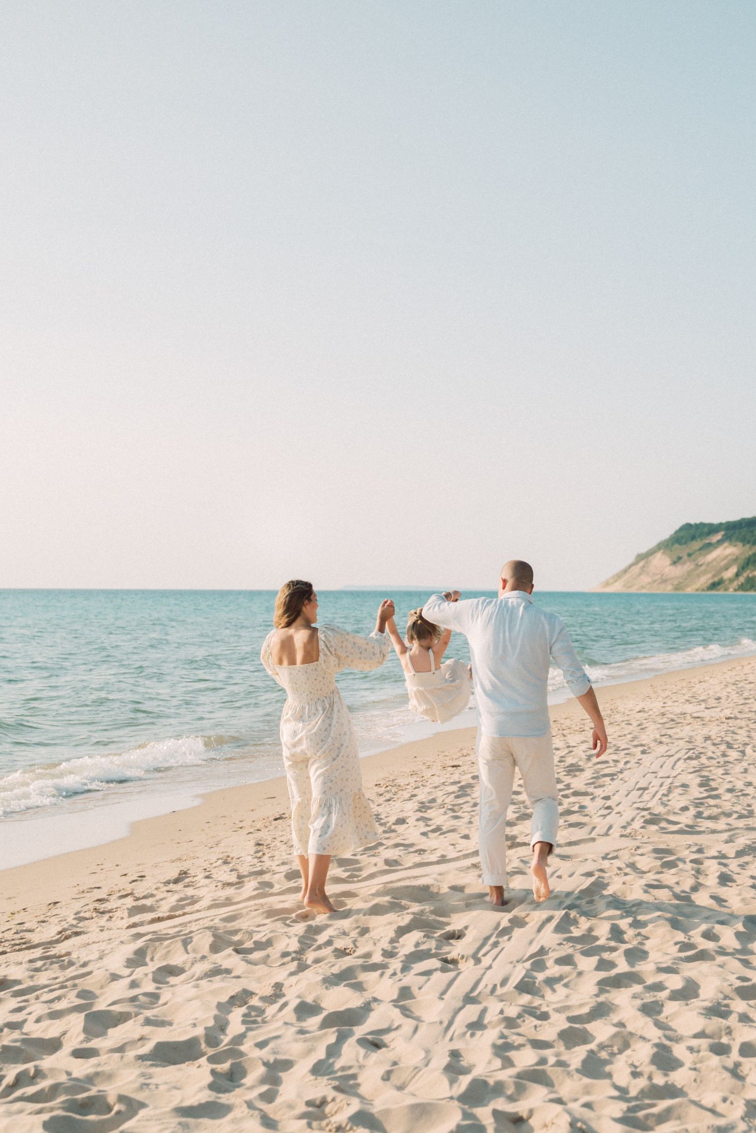 A family in white outfits holds hands while walking along a scenic beach with cliffs in the background.