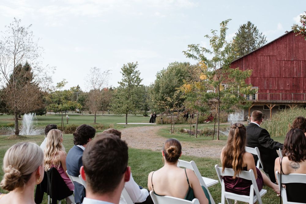 A group of wedding guests seated on white chairs at an outdoor ceremony near a rustic red barn in autumn.
