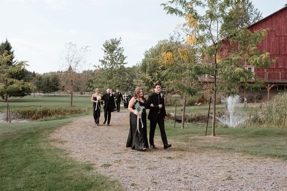 Wedding party members gather on a gravel path near a rustic red barn during an autumn countryside celebration.