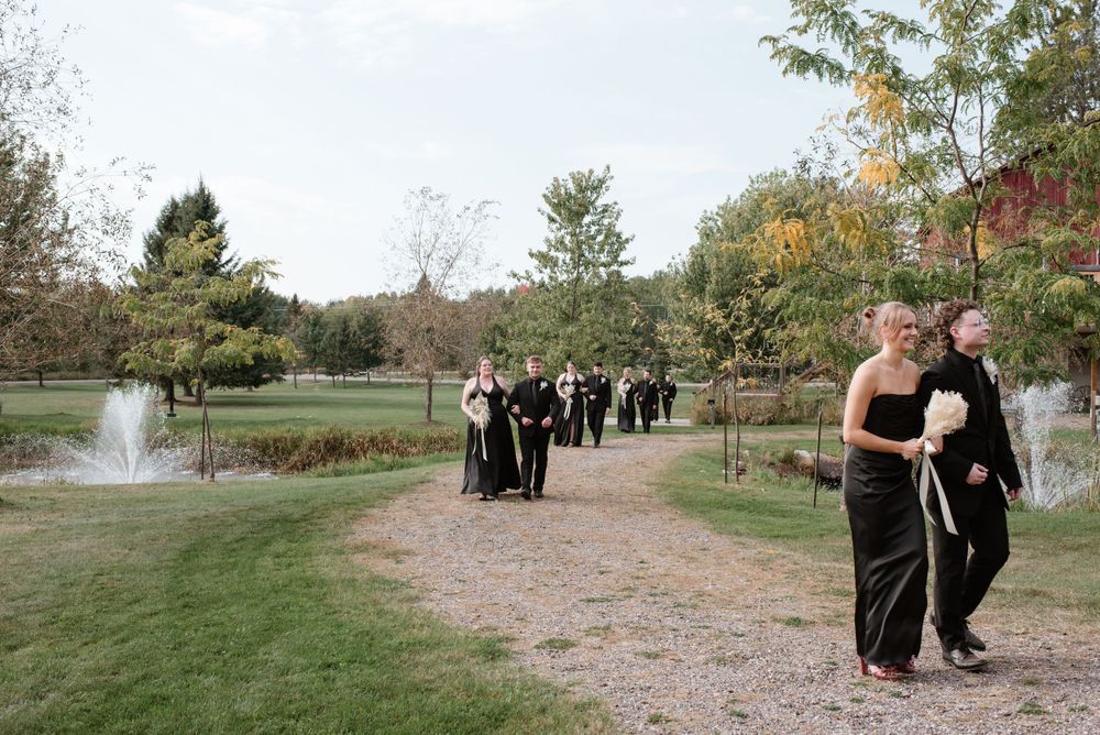 Wedding party members in formal black attire walk along a scenic garden path with a fountain in the background.