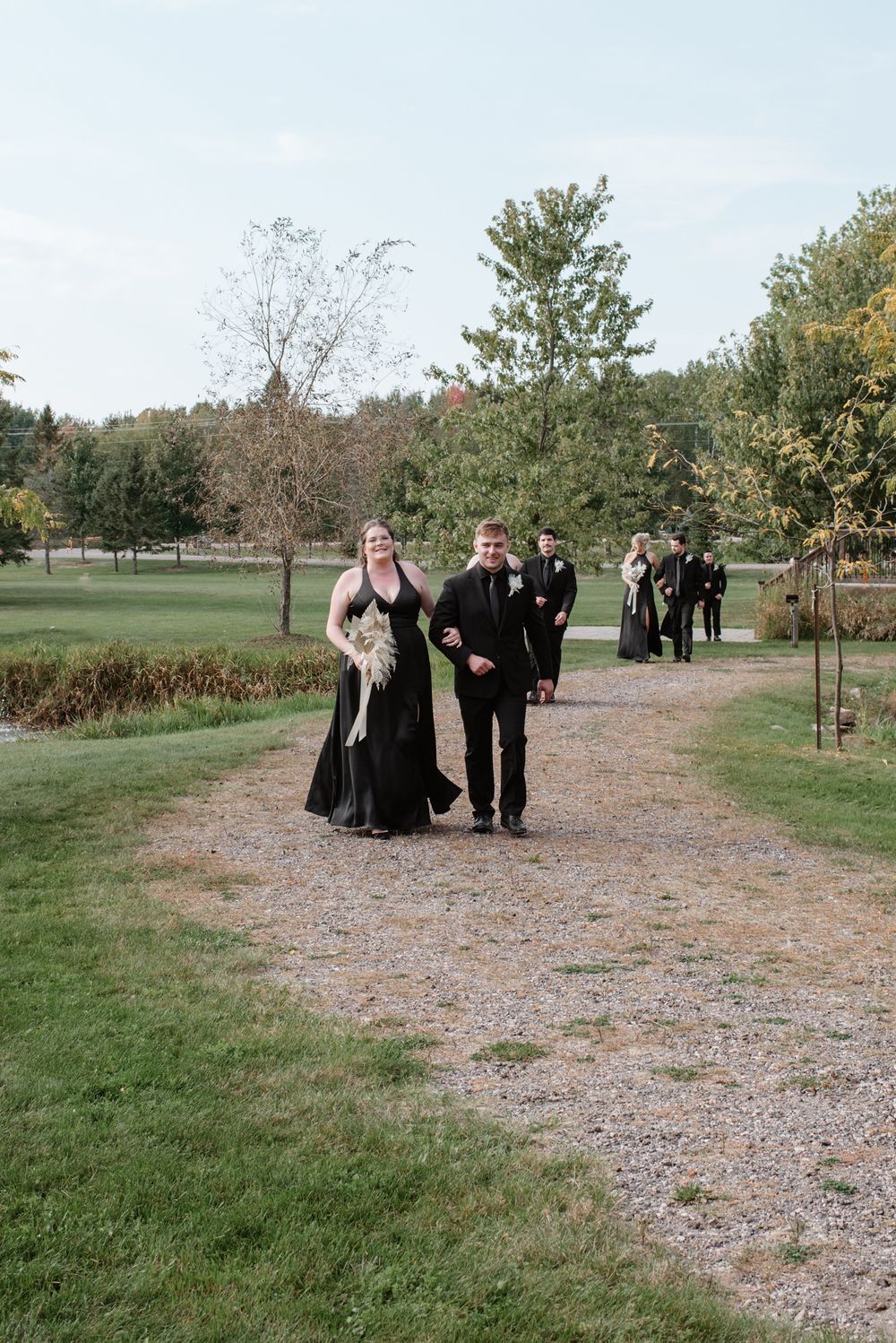 Wedding party members walk along a gravel path through a scenic outdoor garden venue on an autumn day.