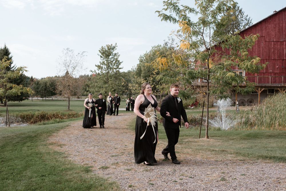 Wedding bridal party walking along a path near a rustic red barn with fall foliage in the background.