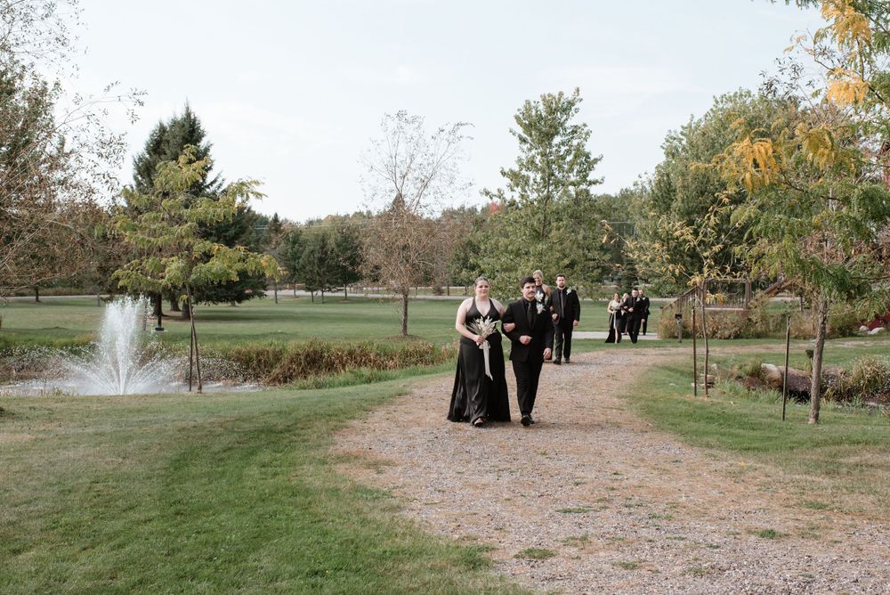 People in formal attire walk along a path in a peaceful park setting with a fountain and autumn trees.