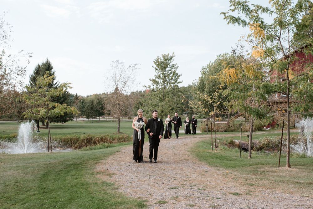 Wedding guests walk along a dirt path in a garden setting with a fountain and autumn trees in the background.