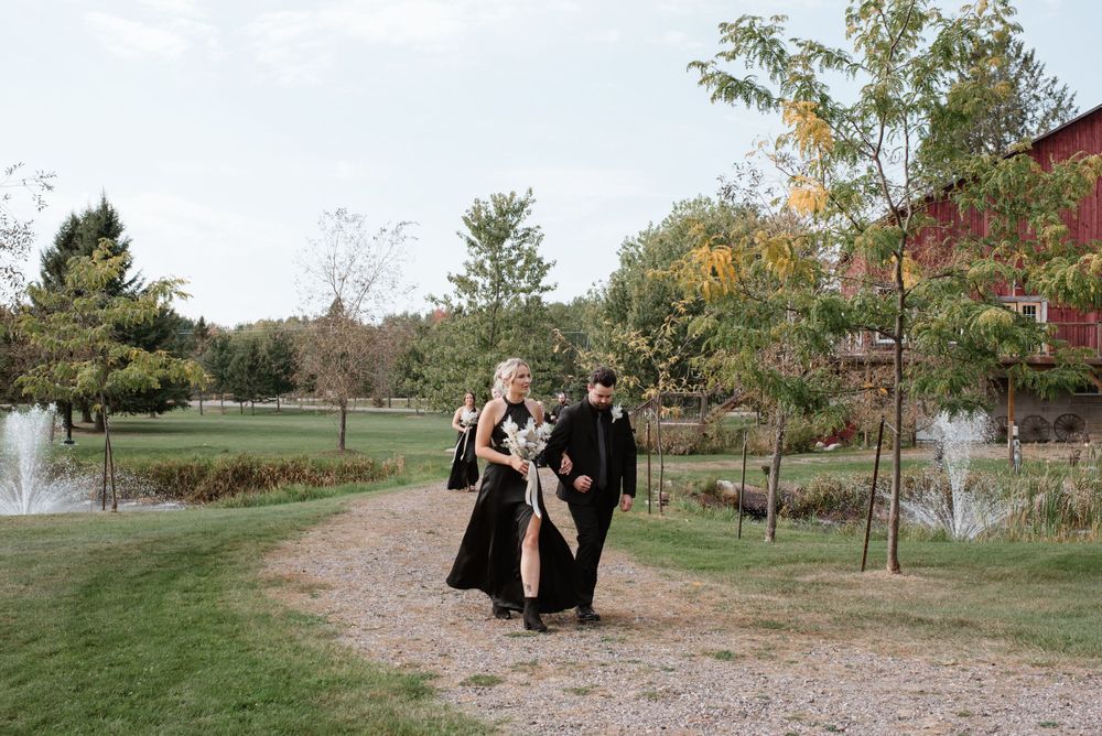 People in dark graduation gowns walk together on a path through a scenic park with fountains and autumn trees.