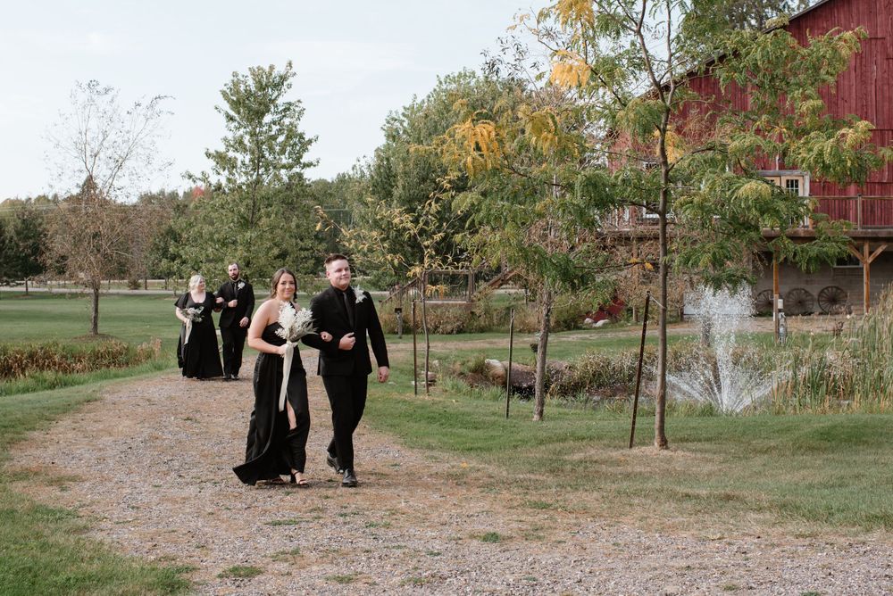 A group dressed in elegant black attire walks along a gravel path next to a red barn on an autumn day.