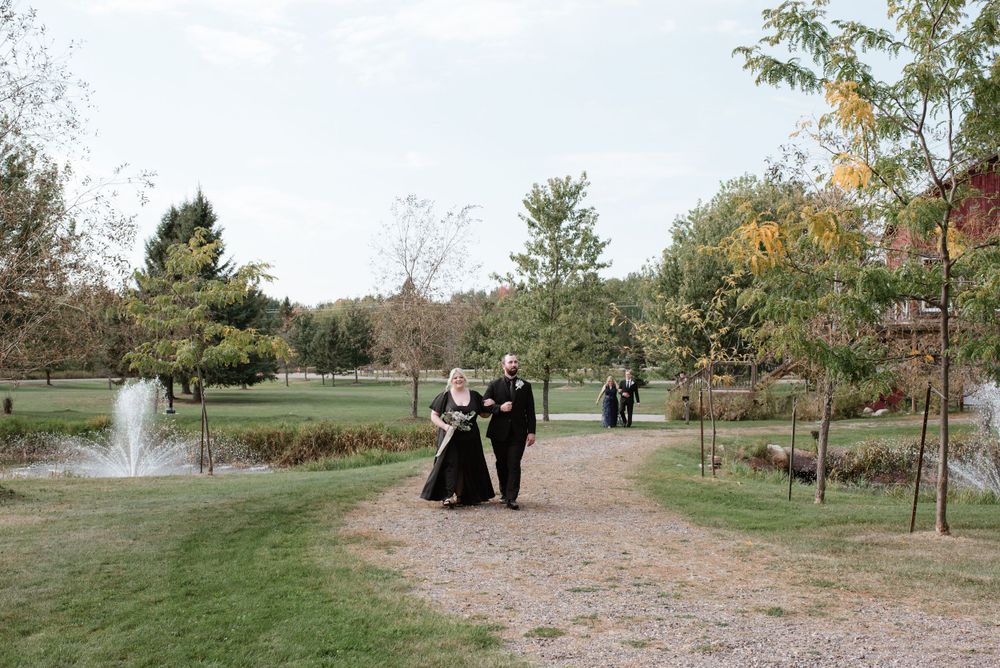 Formal wedding party walking down a gravel path in a picturesque park setting with a fountain and autumn trees.