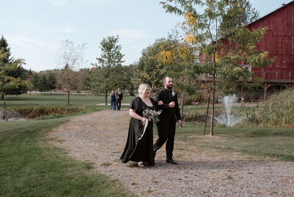 A couple in formal black attire walks along a gravel path near a red barn and pond on an autumn day.