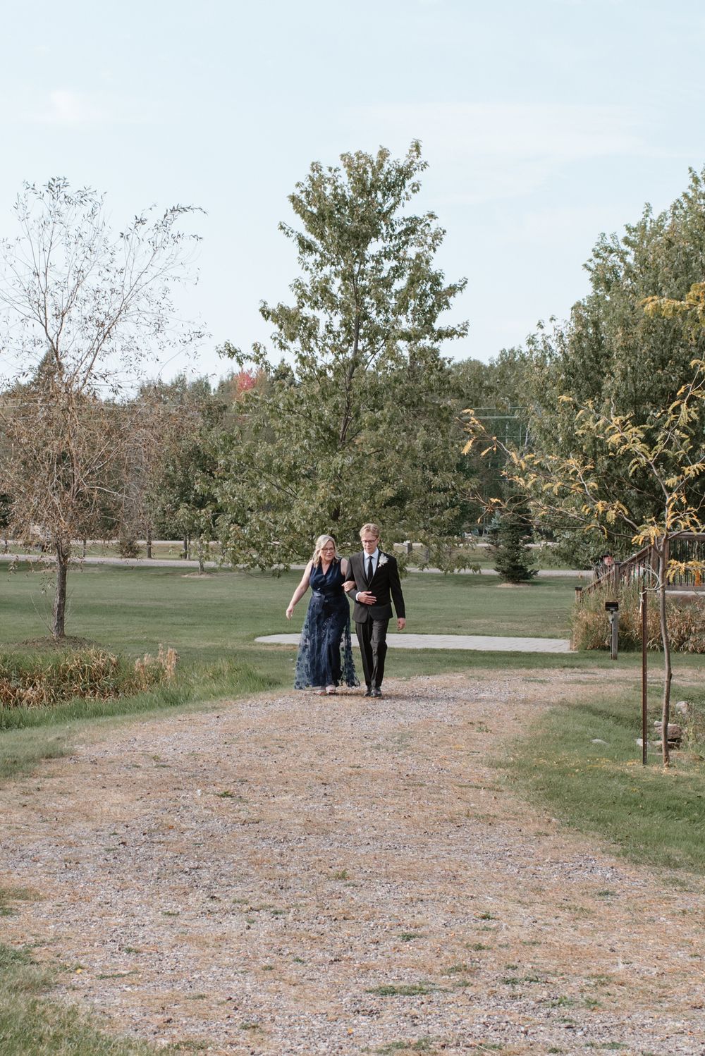 A couple walks together on a gravel path through a serene park landscape with autumn trees and grass.