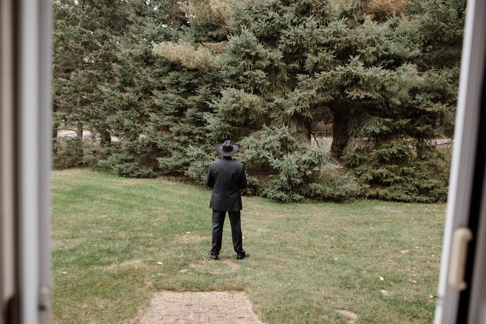 A person in a dark suit stands alone on a grassy lawn surrounded by evergreen trees.