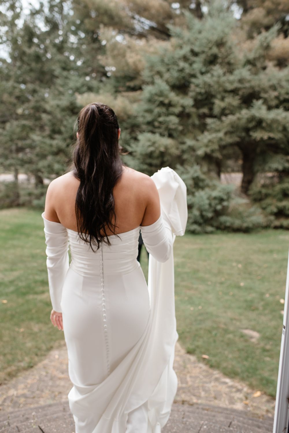 Back view of a bride in an elegant white fitted wedding dress with long sleeves standing on an outdoor path.