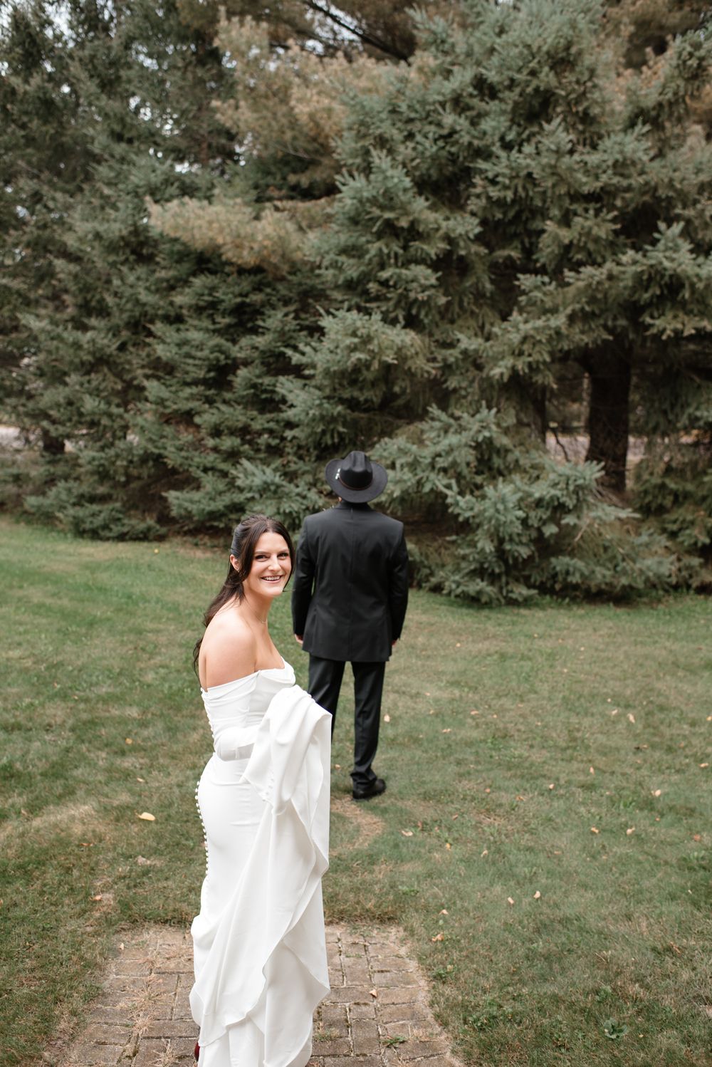 A bride in a white off-shoulder dress glances back while walking on a path through evergreen trees with someone ahead.