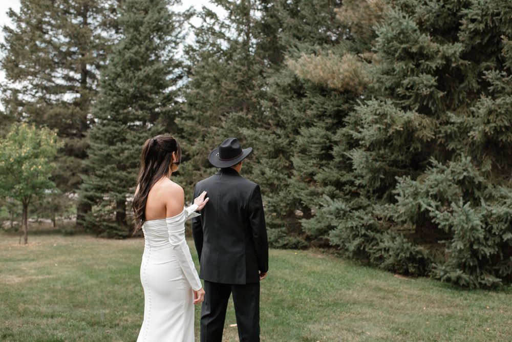 Two people in formal wedding attire stand together looking towards evergreen trees in a grassy outdoor setting.