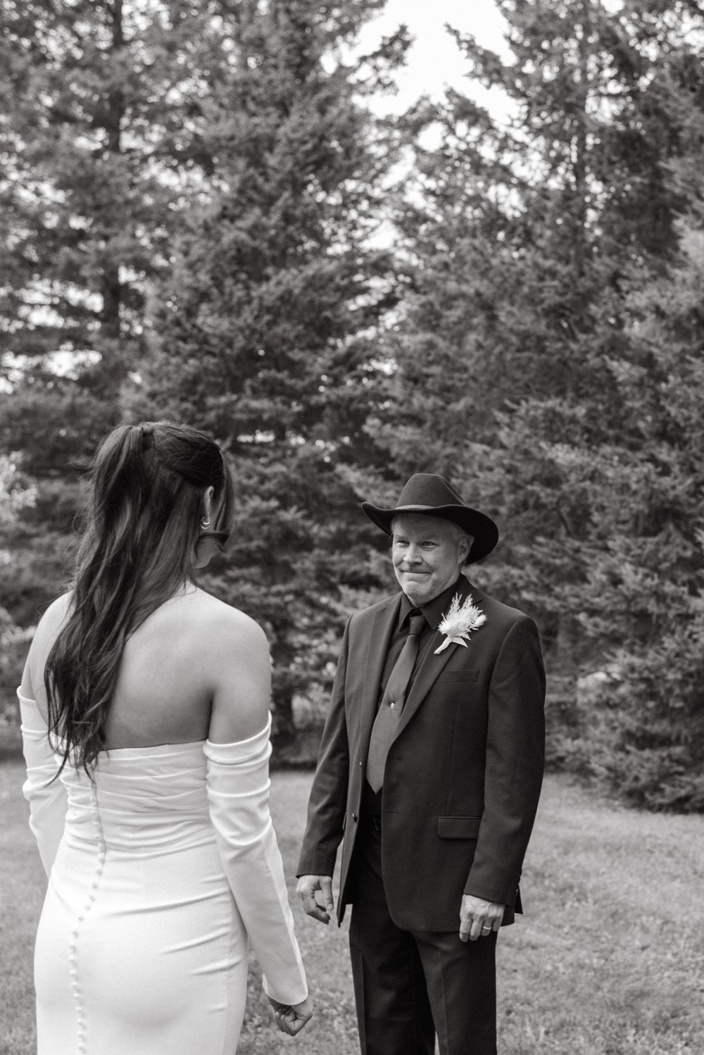 A first look wedding moment between a bride in white dress and wedding guest in suit and hat among pine trees.