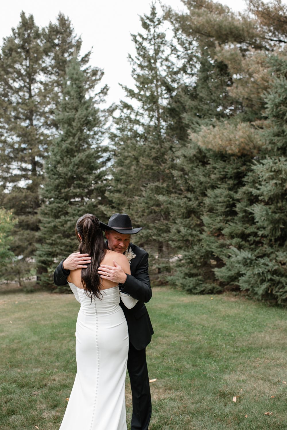 A couple embraces in an intimate moment outdoors with tall evergreen trees creating a romantic forest backdrop.