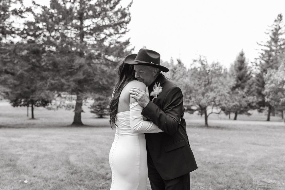 A black and white wedding photo shows a couple sharing a tender embrace on a grassy lawn surrounded by pine trees.