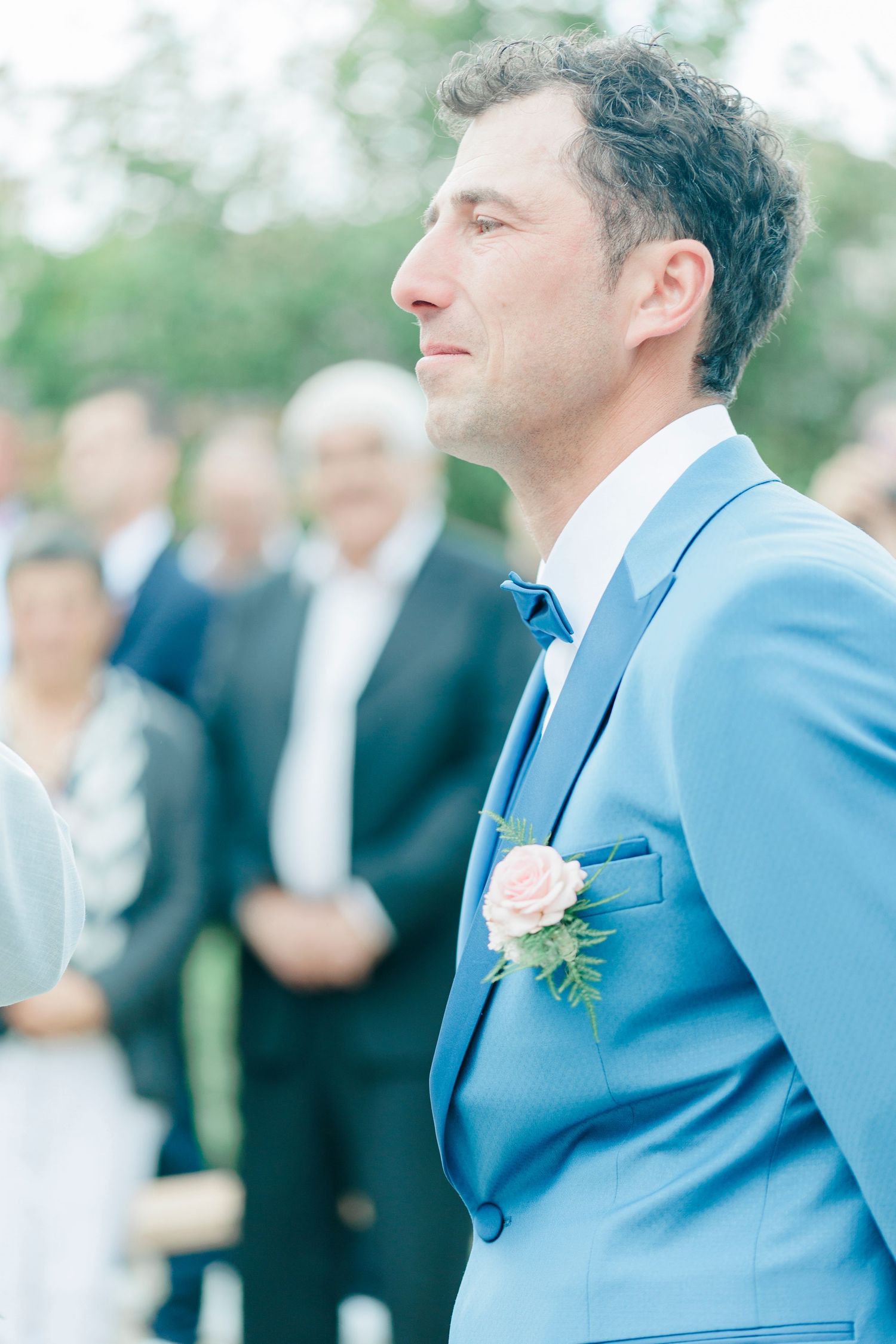 A person in a powder blue suit with pink boutonniere stands during an outdoor ceremony.