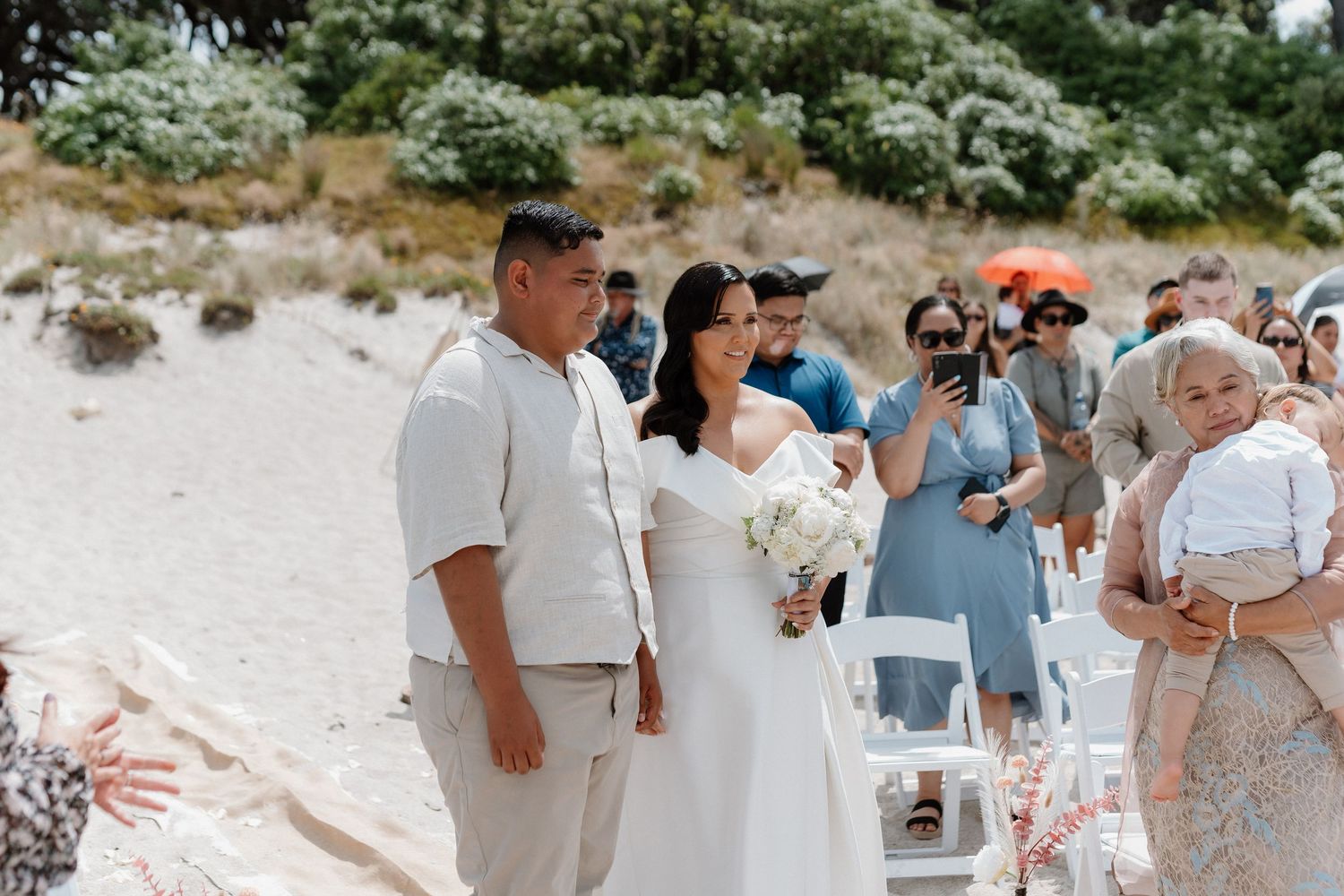 Beachside wedding ceremony with wedding party and guests gathered on sandy shore surrounded by greenery.