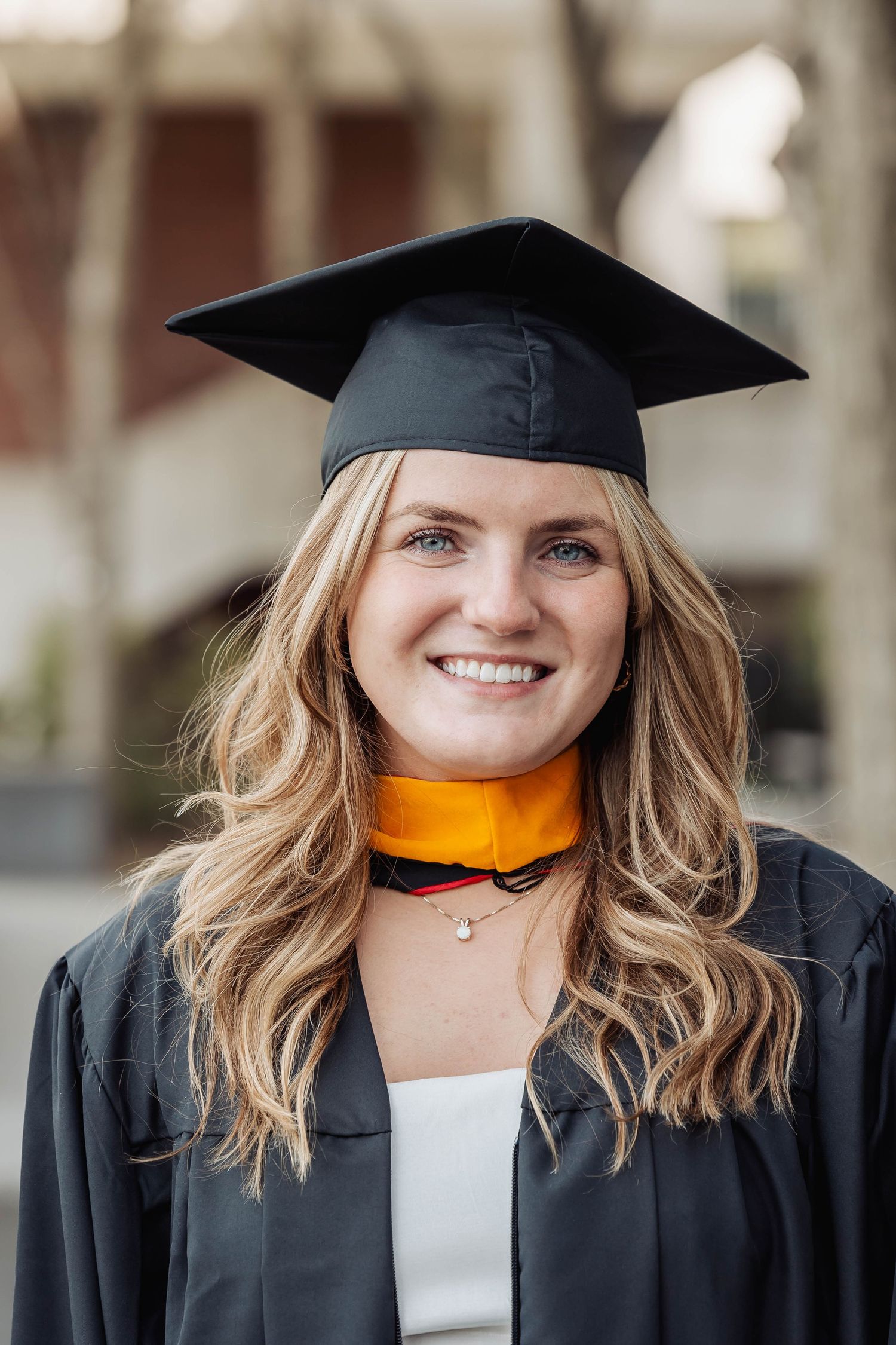 Graduate wearing orange and black commencement regalia with mortarboard cap poses outdoors with blossoming trees in background.