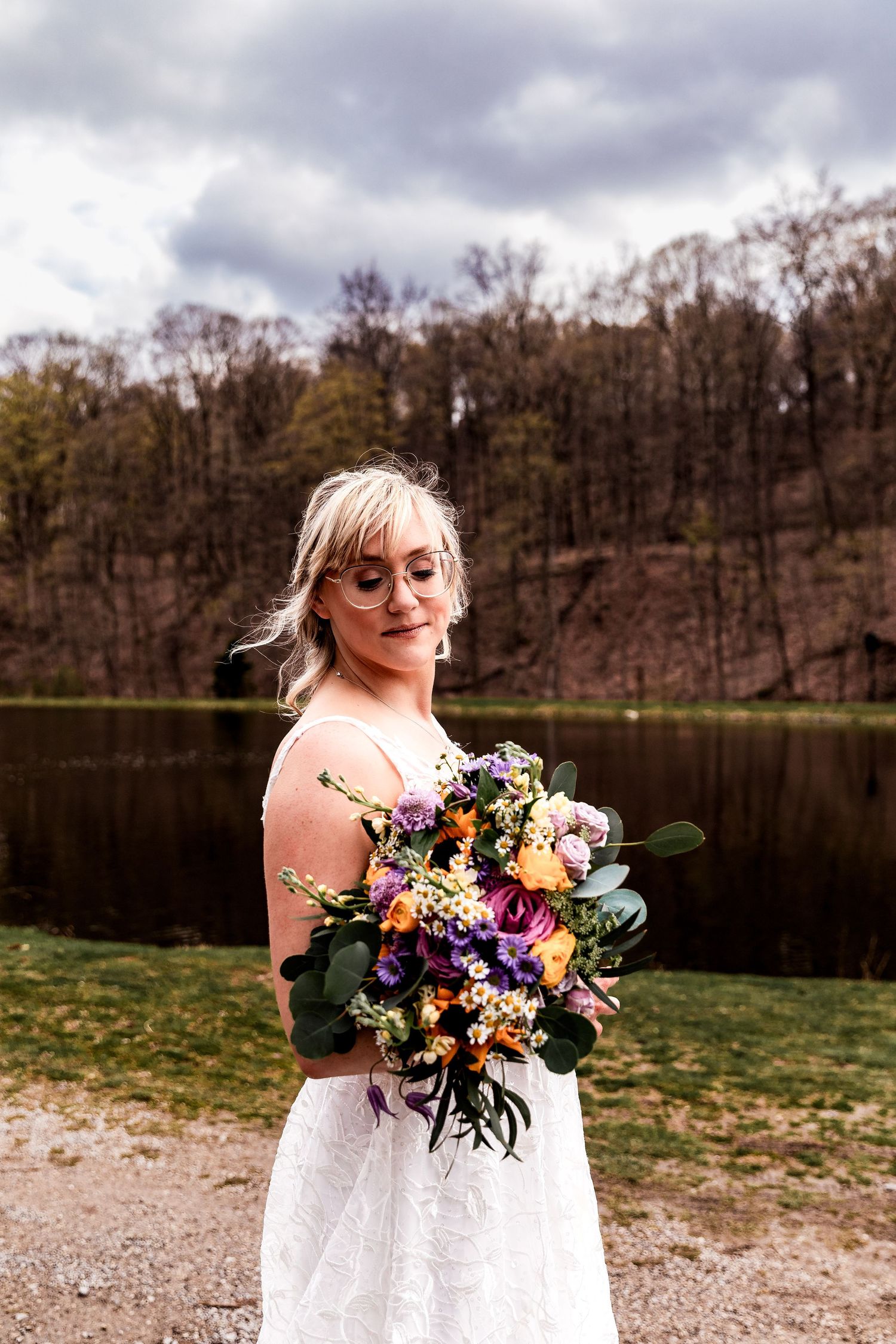 A romantic lakeside scene with a vibrant wildflower bouquet featuring purple, orange and yellow blooms against a cloudy sky.